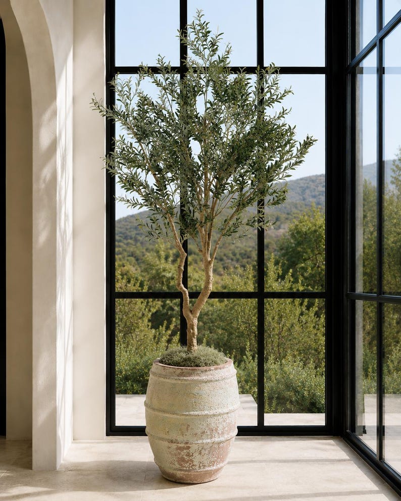 May include: A tall, slender artificial olive tree with green leaves and a textured, light-colored trunk, planted in a large, weathered, barrel-shaped ceramic planter with a distressed, earthy finish and visible horizontal ridges.