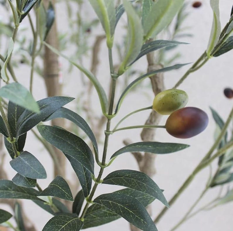 May include: An artificial olive tree branch featuring slender, dark green leaves with visible veins and a textured, light brown stem. The branch includes small, round olives in varying shades of light green and dark purple, attached to thin green stems.