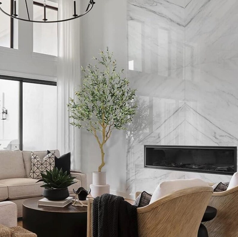 May include: A living room interior with a large artificial olive tree in a white pot, a fireplace with a black surround, and two chairs with white cushions and wooden frames.