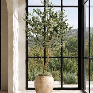 May include: A tall artificial olive tree with slender green leaves and thin branches, planted in a large, round, textured beige ceramic pot with a weathered finish. The tree features a central trunk that splits into multiple stems reaching upward.
