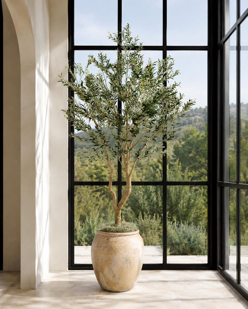 May include: A tall artificial olive tree with slender green leaves and thin branches, planted in a large, round, textured beige ceramic pot with a weathered finish. The tree features a central trunk that splits into multiple stems reaching upward.