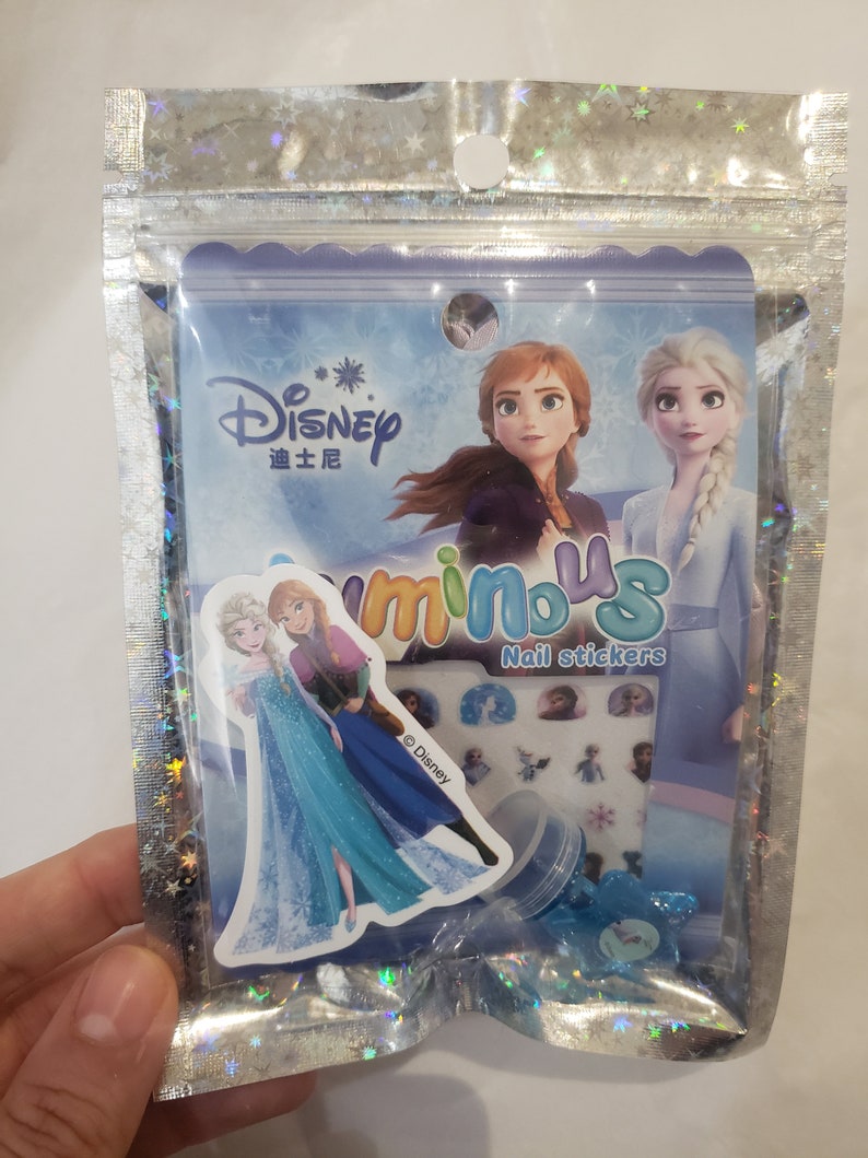 May include: A clear plastic bag with a holographic finish containing a set of Frozen nail stickers. The bag features a colorful image of Elsa and Anna from the Disney movie Frozen. The text "Disney" and "Nail Stickers" are visible on the packaging.
