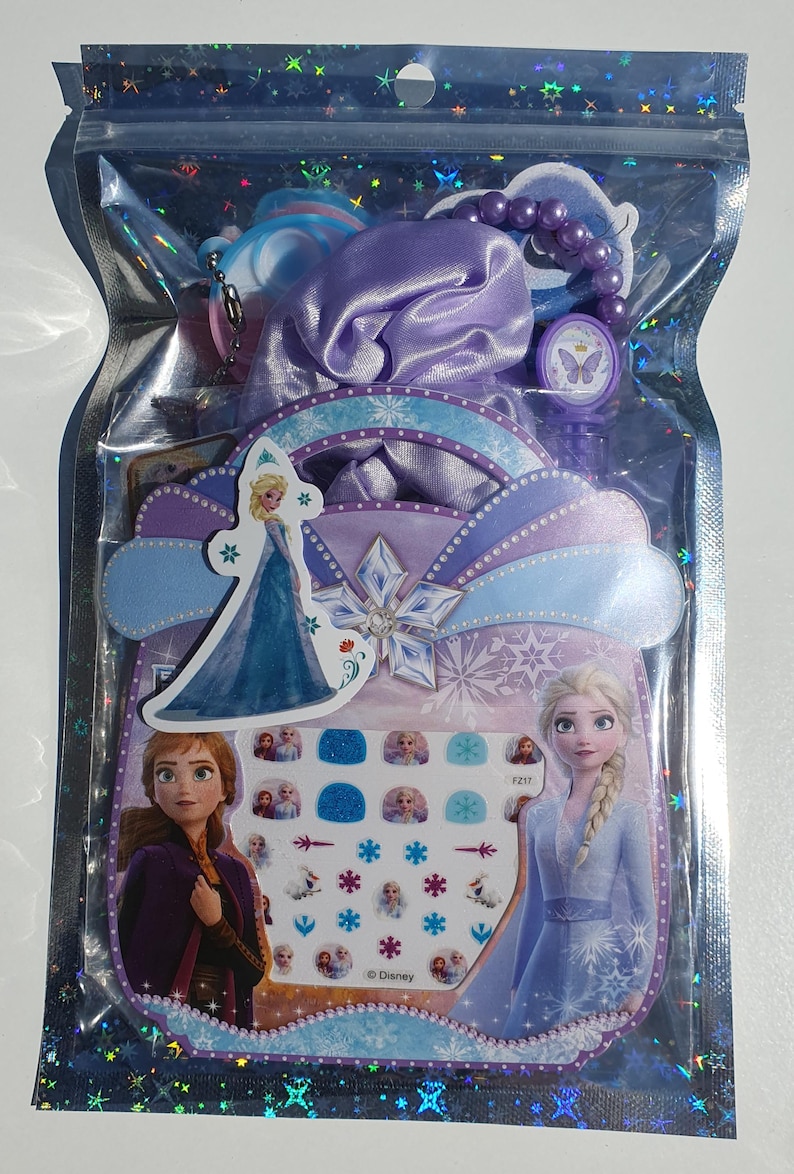 May include: A clear plastic bag filled with Frozen-themed accessories. The bag contains a purple satin hair scrunchie, a purple beaded bracelet, a butterfly-shaped charm, and a sheet of nail stickers featuring images of Elsa and Anna. The nail stickers also include snowflakes and other winter-themed designs. The bag has a white background with a blue and purple border. The text "&copy; Disney" is printed on the bag.