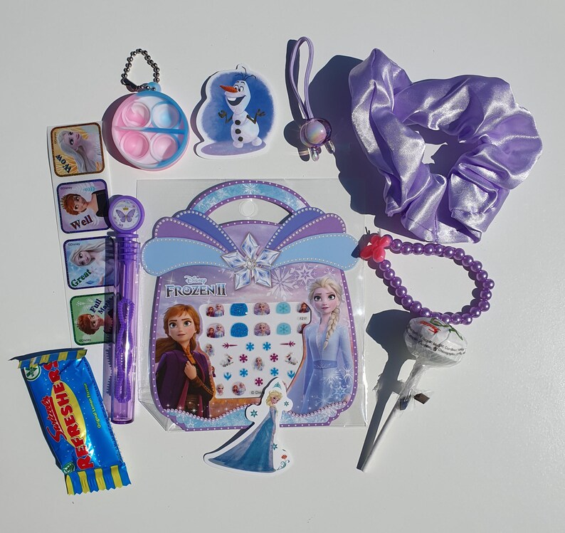 May include: A Frozen II themed gift bag filled with various items, including a purple hair tie, a purple beaded bracelet, a lollipop, a sticker sheet, a small plastic container with a lid, a small plastic toy, a candy bar, and a small plastic toy. The bag is decorated with a picture of Elsa and Anna from Frozen II.