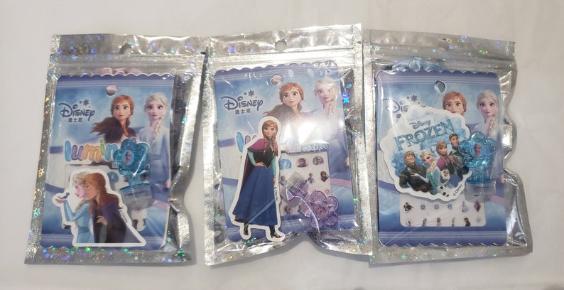 May include: Three plastic bags with different Disney Frozen themed sticker sets. Each bag features a different character from the movie, including Anna, Elsa, and Olaf. The bags are decorated with glitter and have a blue and white color scheme.