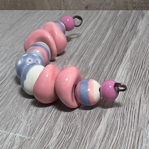 May include: A strand of ceramic beads in shades of pink, white, and blue. The beads have various shapes, including round, donut, and heart-shaped. Some beads feature star patterns or striped designs. Each end has a small metal loop.