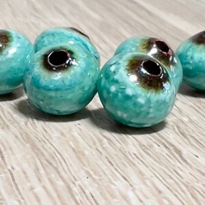 May include: A group of six turquoise and brown ceramic beads with a hole in the center. The beads have a speckled and glazed finish.
