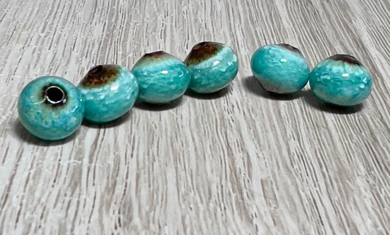 May include: Six teal blue and brown ceramic beads with a glossy finish. The beads are round and have a hole in the center for stringing.