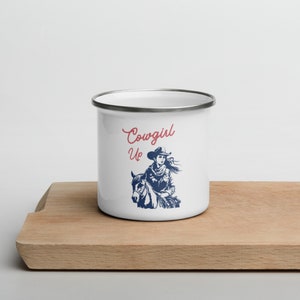 May include: White enamel mug with a blue and white illustration of a cowgirl riding a horse. The text "Cowgirl Up" is written in red on the mug.