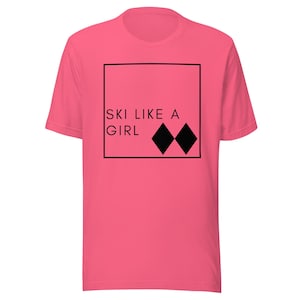 Può includere: Una maglietta rosa con la scritta "SKI LIKE A GIRL" all'interno di un quadrato nero, accompagnata da due forme a diamante nere. La maglietta è realizzata in materiale morbido e ha maniche corte.