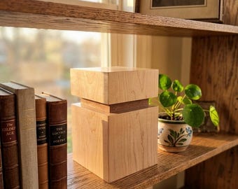 May include: A light-colored wooden box with a stepped design sits on a wooden shelf. The shelf also holds several books, a small potted plant, and a framed portrait. The box is the focal point.