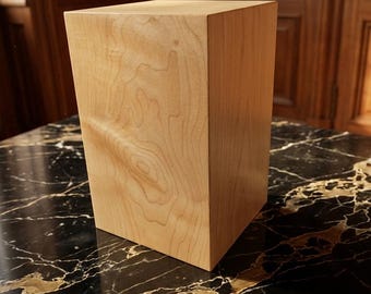 May include: A light-colored wooden block with visible wood grain. The block is rectangular and stands upright on a black and gold marble surface. The wood has a natural, warm tone.
