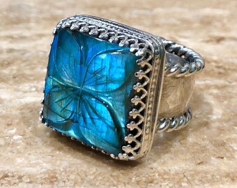 May include: A silver ring with a square blue gemstone set in a decorative silver frame. The gemstone has a carved floral design.
