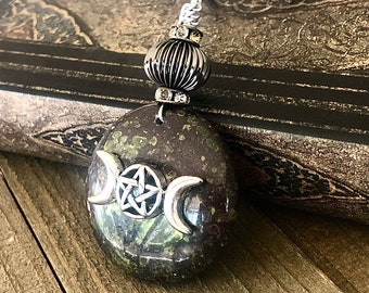 Può includere: Una collana con catena d'argento con un ciondolo in pietra verde e marrone con un design a tripla luna e pentacolo. Il ciondolo è attaccato a una perla d'argento con strass.