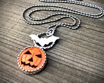 May include: A silver chain necklace with a carved orange pumpkin charm and a silver bat charm.