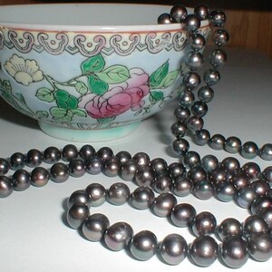 Pu&ograve; includere: Una collana di perle grigio scuro drappeggiata su una ciotola di porcellana blu e bianca con motivi floreali. Le perle sono rotonde e lisce, e la ciotola presenta un delicato motivo floreale.