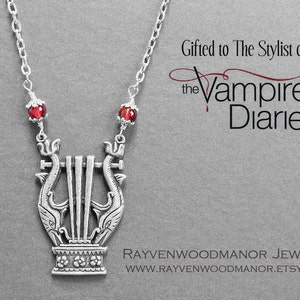 May include: Silver necklace with a lyre pendant and red glass beads. The pendant features two dolphins flanking the lyre. The necklace is labeled "Gifted to The Stylist of the Vampire Diaries".