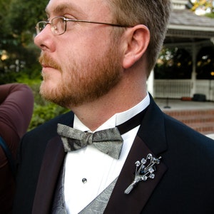 May include: A man wearing a black tuxedo with a white shirt and a silver bow tie. He is wearing a silver pin with crystals on his lapel.