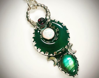 May include: A silver necklace with a green jade pendant featuring a white opal, a red garnet, and a green labradorite gemstone. The pendant is shaped like a crescent moon with intricate silver details.