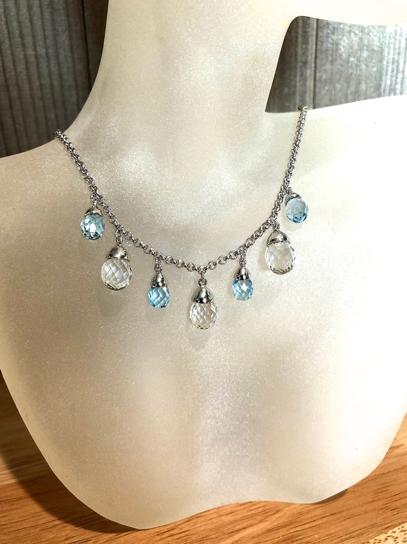 May include: A silver chain necklace with clear and light blue faceted gemstone drops. The necklace is displayed on a white mannequin bust.