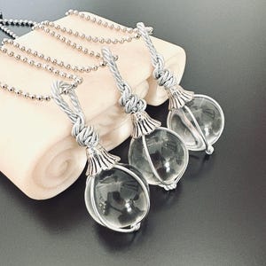 May include: Three clear glass orb pendants with silver-colored metal accents and chains. Each orb is encased in a wire frame and suspended from a twisted metal loop. The pendants are displayed on a cream-colored surface against a dark background.