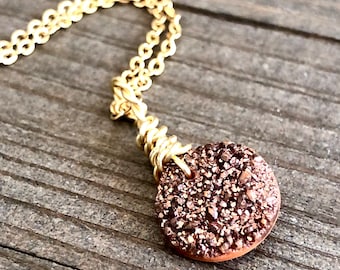 Può includere: Una collana con catena dorata con un ciondolo rotondo color rame. Il ciondolo è fatto di piccoli cristalli scintillanti.