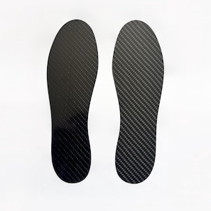 Op de afbeelding: Een paar zwarte schoenzolen met een opvallend koolstofvezel weefpatroon. De inlegzolen hebben een gladde, langwerpige en voorgevormde vorm die is ontworpen om in schoenen te passen, met een donker, reflecterend en gestructureerd oppervlak.