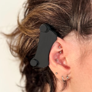 以下が含まれることがあります： 2つの小さな黒いボタンが付いた黒いプラスチック製のイヤホン。イヤホンは耳に装着し、聴覚を助けるように設計されています。