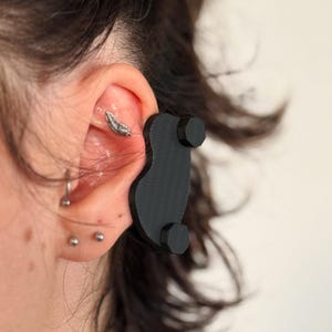 May include: A black 3D printed ear piece designed to hold a small device, such as a hearing aid, in place. The ear piece has two small, hexagonal-shaped protrusions on the bottom.