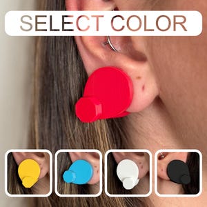May include: Red earplugs with a circular design and a small, cylindrical handle. The image shows the earplugs in use, with additional color options: yellow, blue, white, and black. The text "SELECT COLOR" is displayed at the top.