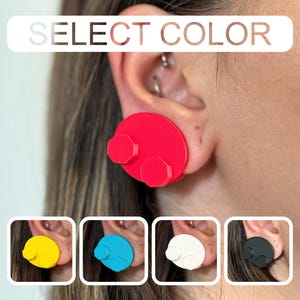 May include: Red, round earrings with two hexagonal accents. The image shows the earrings on an ear, with additional color options in yellow, blue, white, and black. The text "SELECT COLOR" is displayed at the top.