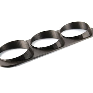 Puede incluir: Un objeto de plástico negro impreso en 3D con tres recortes circulares. El objeto está diseñado para contener tres objetos redondos, como medidores u otros objetos pequeños.
