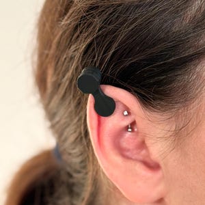 May include: A black ear clip, designed to fit over the ear. The clip has a dumbbell-like shape with a rounded top and a curved bottom section. The ear clip is shown on a person's ear.