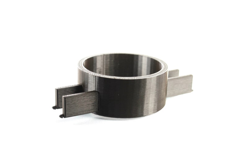 K&ouml;nnte beinhalten: Ein schwarzer 3D-gedruckter Ring mit zwei Armen, die sich zu den Seiten erstrecken. Der Ring ist so konzipiert, dass er etwas an Ort und Stelle h&auml;lt.