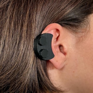 Puede incluir: Un auricular negro con dos pequeños botones negros en el lateral. El auricular se lleva en la oreja de una persona con cabello castaño.