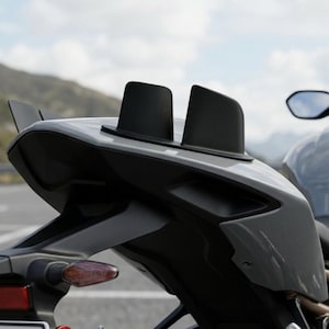 May include: The rear section of a gray motorcycle featuring two prominent black vertical fins mounted on the tail fairing. The design includes angular bodywork, a red taillight, and a side-mounted turn signal indicator with an orange lens.