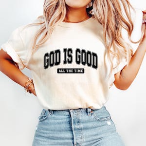 Puede incluir: Camiseta color crema con la frase "GOD IS GOOD" en negro, con una fuente de estilo universitario, y "ALL THE TIME" debajo, en negro más pequeño. La camiseta se combina con unos vaqueros azules.