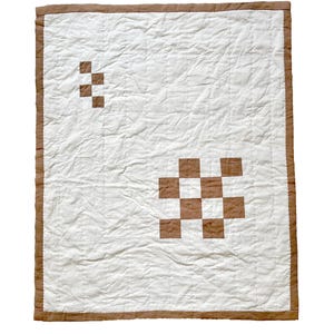 May include: A white quilt with a brown border and two brown checkerboard patterns. The quilt has a quilted texture and a simple, minimalist design. The brown border frames the white center, creating a clean and modern aesthetic.