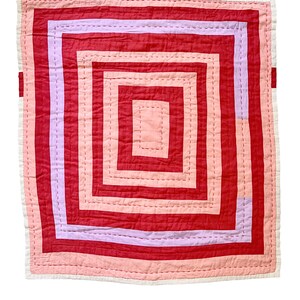 May include: A square, handmade quilt with a concentric square design. The quilt features a color palette of red, pink, and lavender hues. The edges are finished with a white border. The quilt is likely used for bedding or as a decorative item.