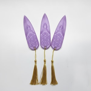 May include: Three purple resin bookmarks with a decorative pattern and gold tassels. The bookmarks are elongated with a pointed tip. The tassels are made of a gold-colored thread. The background is white.