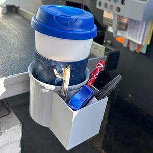 May include: A white cup with a blue lid sits in a gray cup holder. The cup holder has a compartment for pens and pencils. A red pen, a black pen, and a blue pen are in the compartment.