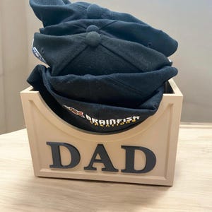 Puede incluir: Una caja de madera beige con la palabra "DAD" en letras negras en la parte delantera. La caja tiene una abertura curva y está llena de gorras de béisbol negras.