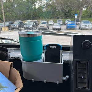 May include: A white 3D printed cup holder with a teal travel mug and a blue USPS device inside. The holder has two hooks on the side.