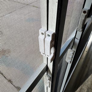 May include: Close-up of a white plastic door latch with a black metal frame. The latch is attached to a screen door with a black mesh.