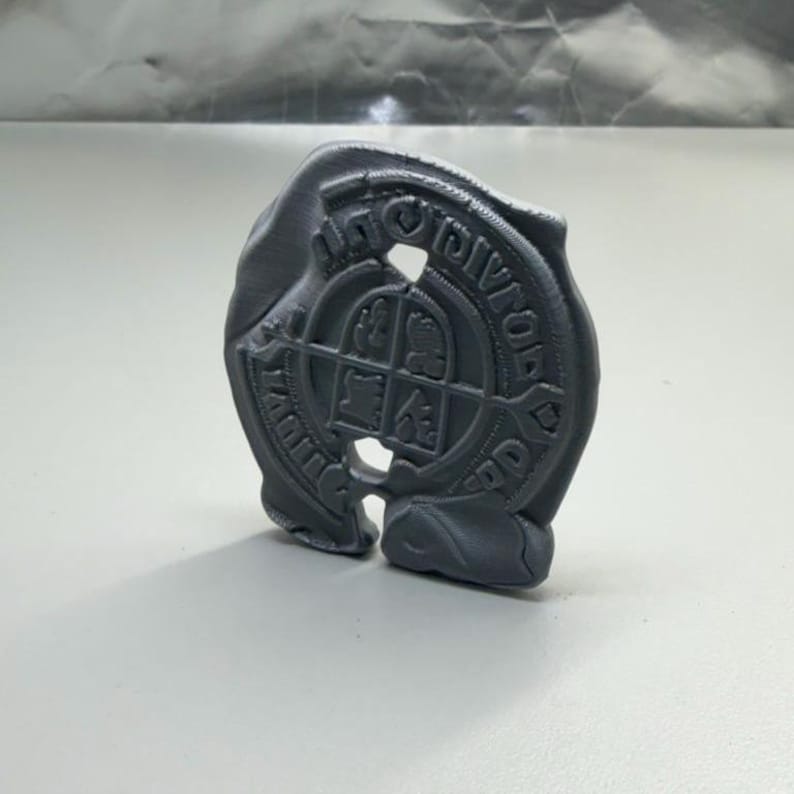 以下が含まれることがあります： 「ROYAL DIVISIONS」の文字と盾のデザインが施された、グレーの3Dプリントエンブレム。エンブレムは円形で、切り欠き部分とテクスチャ加工された表面があります。盾には4つの異なるシンボルが描かれています。