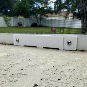 May include: A white rectangular screen frame component with two silver screws. The component is mounted on a concrete surface, with a background of a screen, trees, and a house.