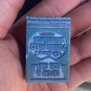 May include: A rectangular, teal-colored printing block with engraved text. The block features two oval text areas and a triangular area with text. The text is in a foreign language. The block is held in a hand.