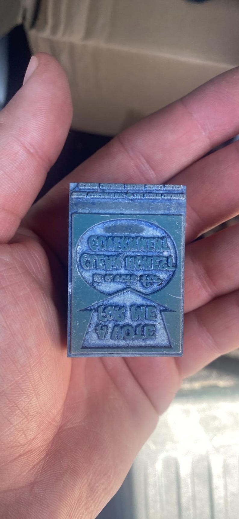 May include: A rectangular, teal-colored printing block with engraved text. The block features two oval text areas and a triangular area with text. The text is in a foreign language. The block is held in a hand.