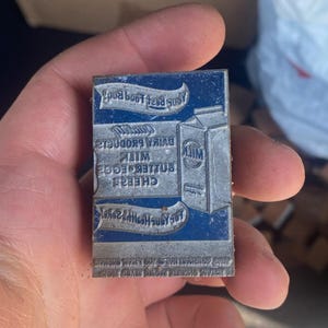 May include: A vintage metal printing block with a blue background and silver text. The text includes the words "Milk", "Butter", "Eggs", and "Cheese". The block is held in a hand.