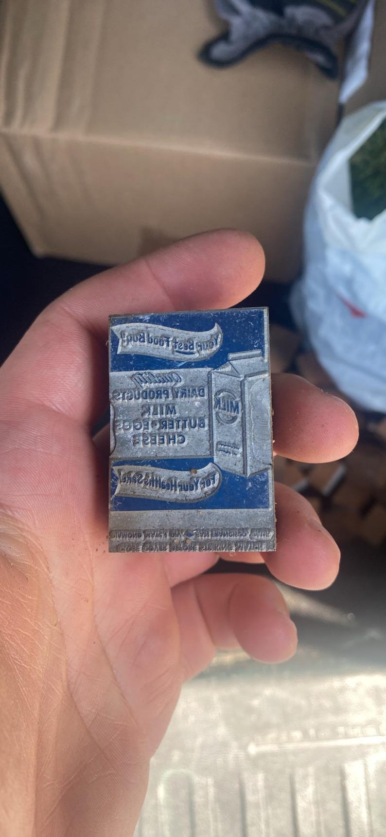 May include: A vintage metal printing block with a blue background and silver text. The text includes the words "Milk", "Butter", "Eggs", and "Cheese". The block is held in a hand.